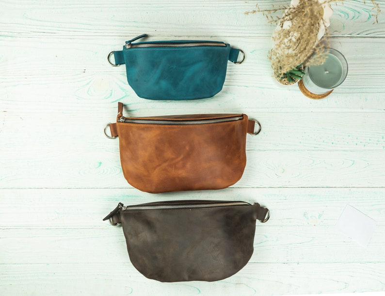 K&ouml;nnte beinhalten: Drei Leder-Bauchtaschen in Teal, Braun und Dunkelbraun. Jede Tasche hat einen Rei&szlig;verschluss und silberfarbene Metallbeschl&auml;ge. Die Taschen sind auf einer hellblauen Holzoberfl&auml;che angeordnet.