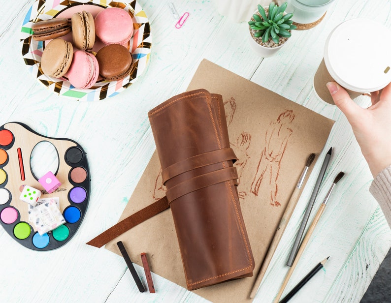 Puede incluir: Un rollo de l&aacute;pices de cuero marr&oacute;n con cierre de correa. El rollo est&aacute; abierto y contiene l&aacute;pices y pinceles. El rollo est&aacute; sobre una mesa con una paleta de acuarela, macarons y una taza de caf&eacute;.