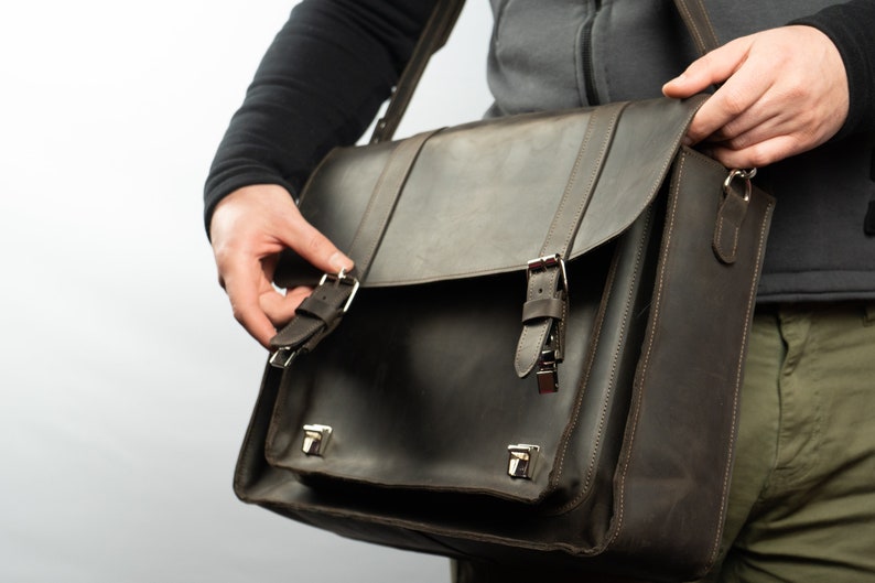 Peut inclure: Un sac messager en cuir brun avec deux boucles et une fermeture &agrave; rabat. Le sac a une bandouli&egrave;re et se porte sur l'&eacute;paule.