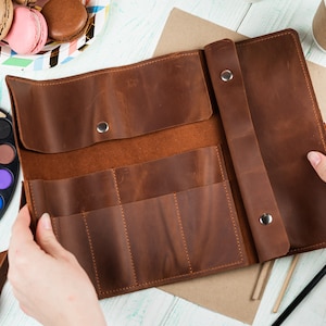 Puede incluir: Un estuche de l&aacute;pices de cuero marr&oacute;n con m&uacute;ltiples ranuras para guardar l&aacute;pices u otros materiales de arte. El estuche est&aacute; abierto y el interior es visible.