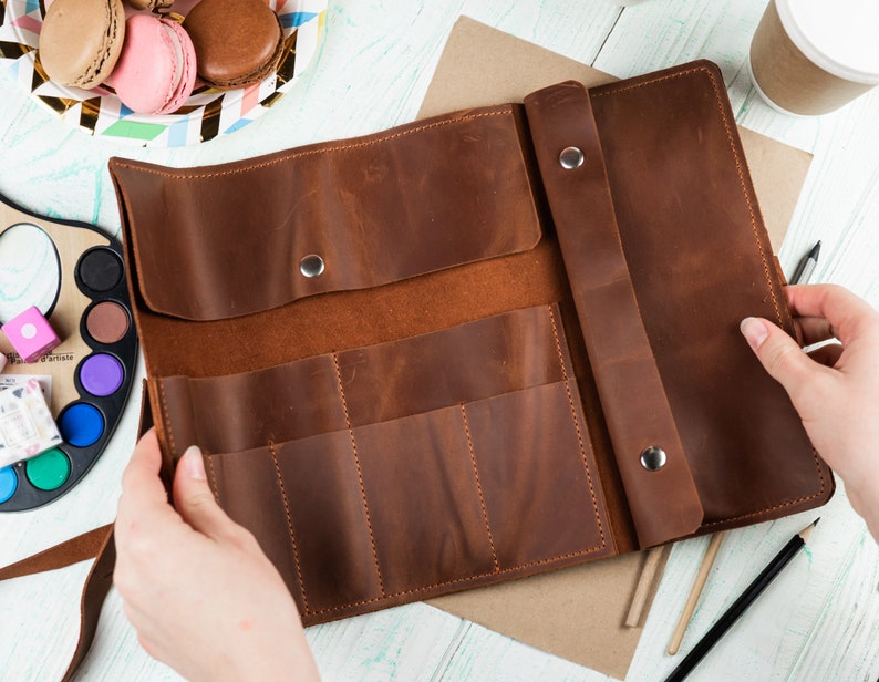 Puede incluir: Un estuche de l&aacute;pices de cuero marr&oacute;n con m&uacute;ltiples ranuras para guardar l&aacute;pices u otros materiales de arte. El estuche est&aacute; abierto y el interior es visible.