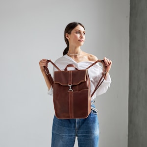 Puede incluir: Una mochila de cuero marr&oacute;n con cierre de solapa y correas ajustables. La mochila tiene un bolsillo frontal y un asa superior. El cuero tiene un aspecto envejecido. Dise&ntilde;ada para llevar libros, port&aacute;tiles y otros elementos esenciales.
