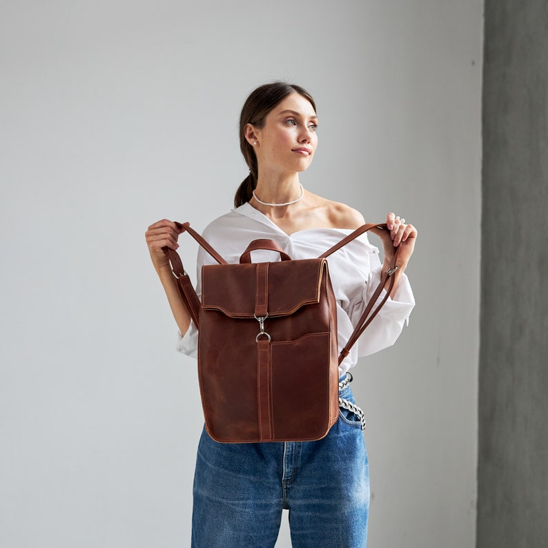 Puede incluir: Una mochila de cuero marr&oacute;n con cierre de solapa y correas ajustables. La mochila tiene un bolsillo frontal y un asa superior. El cuero tiene un aspecto envejecido. Dise&ntilde;ada para llevar libros, port&aacute;tiles y otros elementos esenciales.