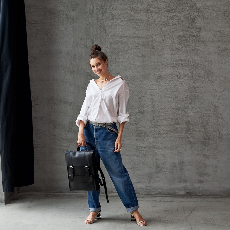 Op de afbeelding: Een vrouw draagt een wit overhemd met knopen, een blauwe jeans en een zwarte leren rugzak. Ze staat voor een grijze betonnen muur.