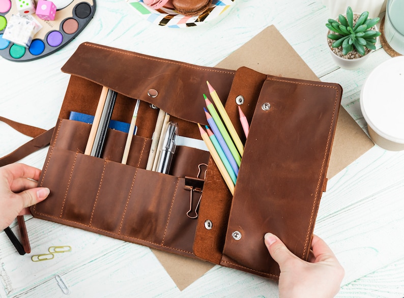 Puede incluir: Un rollo de l&aacute;pices de cuero marr&oacute;n con m&uacute;ltiples ranuras para l&aacute;pices, bol&iacute;grafos y otros materiales de arte. El rollo est&aacute; abierto y tiene una solapa con broches. El rollo est&aacute; sobre una superficie de madera blanca.