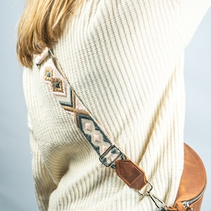 K&ouml;nnte beinhalten: Eine braune Leder-Bauchtasche mit einem gemusterten Riemen mit rosa, blauen und beigen geometrischen Designs. Der Riemen ist mit einem silbernen Verschluss und braunen Lederakzenten befestigt. Die Bauchtasche hat einen Rei&szlig;verschluss.