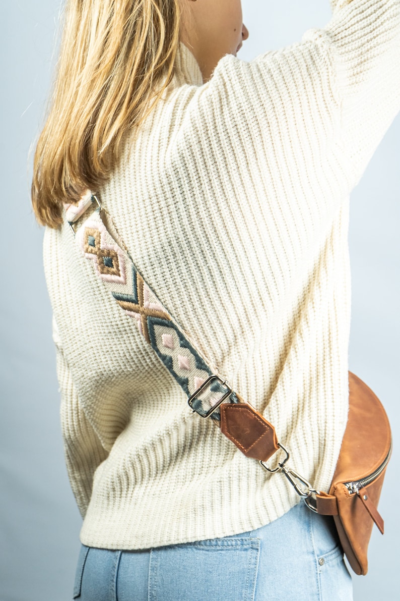 K&ouml;nnte beinhalten: Eine braune Leder-Bauchtasche mit einem gemusterten Riemen mit rosa, blauen und beigen geometrischen Designs. Der Riemen ist mit einem silbernen Verschluss und braunen Lederakzenten befestigt. Die Bauchtasche hat einen Rei&szlig;verschluss.