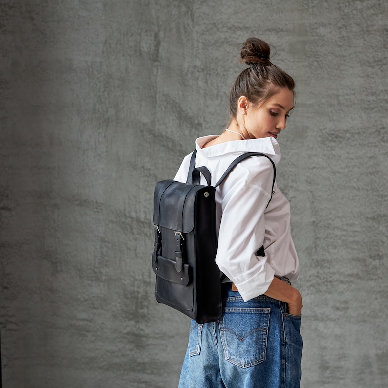 Op de afbeelding: Een zwarte leren rugzak met verstelbare banden en gespen. De rugzak wordt gedragen door een persoon met een wit shirt en een blauwe jeans.