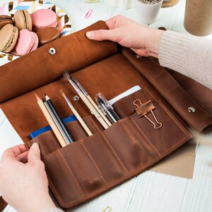 Puede incluir: Un estuche enrollable de cuero marr&oacute;n con m&uacute;ltiples ranuras para guardar art&iacute;culos de arte, como l&aacute;pices, bol&iacute;grafos y pinceles. El estuche est&aacute; abierto y las ranuras son visibles.