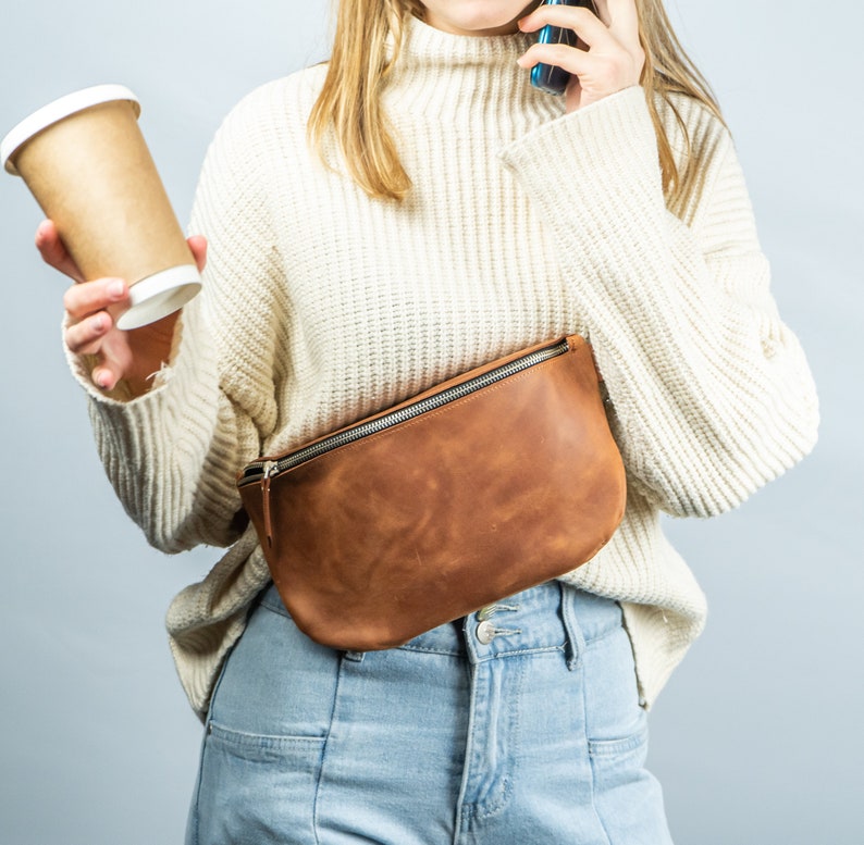 K&ouml;nnte beinhalten: Eine braune Leder-Bauchtasche mit silbernem Rei&szlig;verschluss wird &uuml;ber hellblauen Jeans getragen. Eine Person h&auml;lt eine Kaffeetasse und ein Telefon. Die Person tr&auml;gt einen cremefarbenen Pullover.