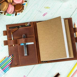 May include: A brown leather journal with a zipper closure and multiple pockets. The journal is open to reveal blank pages and a pen in one of the pockets. There are also colored pencils and a pencil in the image.