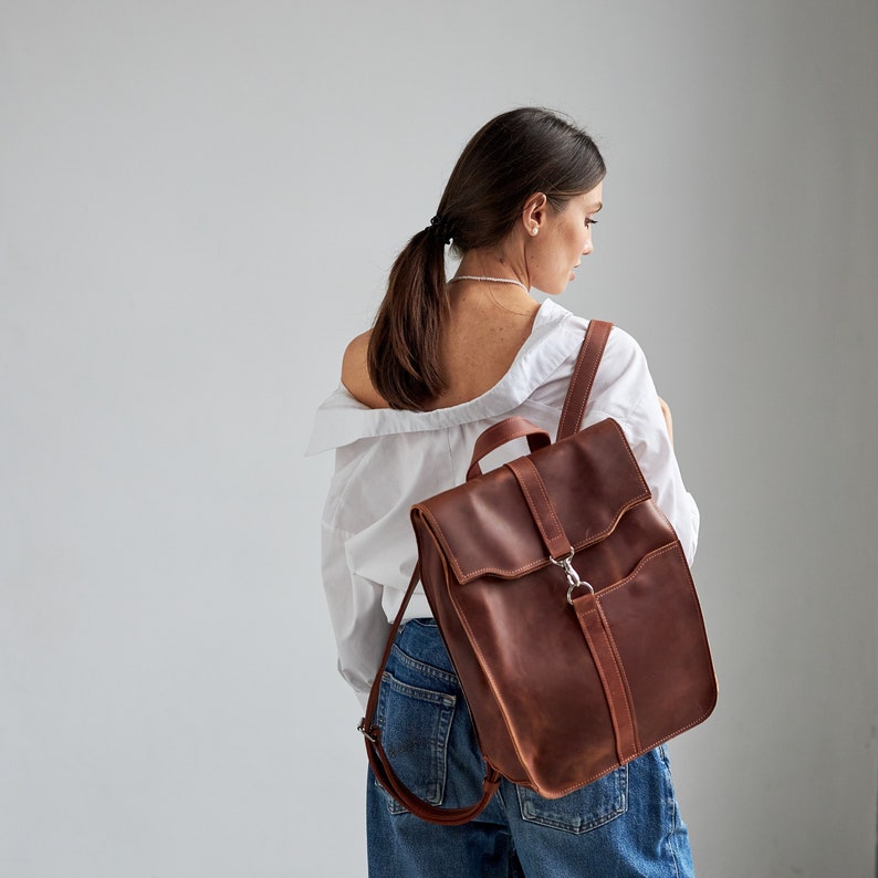 Puede incluir: Una mochila de cuero marr&oacute;n con cierre de solapa y correas ajustables. La mochila tiene un asa en la parte superior y un cierre plateado. La bolsa se lleva sobre el hombro.
