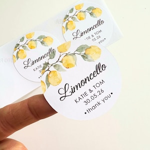 Peut inclure: Ronds autocollants blancs avec le mot "Limoncello" en écriture cursive, ornés de citrons et de feuilles à l'aquarelle. Les autocollants incluent des noms et une date, parfaits pour un mariage ou un événement.