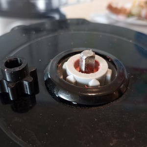 Peut inclure: Gros plan sur une base de robot culinaire noire avec une broche centrale et un engrenage noir détaché. La broche a un poteau métallique entouré d'un composant en plastique blanc et rouge. L'engrenage est en forme d'étoile.