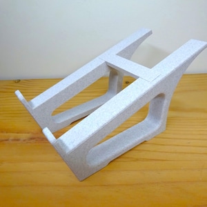 Puede incluir: Un soporte para portátil gris claro con superficie texturizada. El soporte tiene un diseño abierto con soportes angulados y una base horizontal. Diseñado para sostener un portátil y elevarlo.