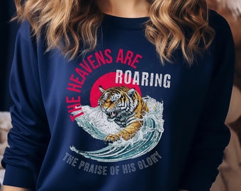 Puede incluir: Una sudadera azul marino con un diseño gráfico de un tigre rugiendo en una ola. El texto "The Heavens Are Roaring The Praise Of His Glory" está impreso en la sudadera.