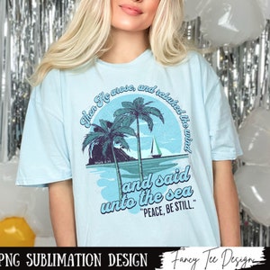Op de afbeelding: Een lichtblauw t-shirt met een grafisch ontwerp van palmbomen, een zeilboot en een blauwe cirkel met de tekst "Then He arose, and rebuked the wind Mark 4:39 and said unto the sea "PEACE, BE STILL.""