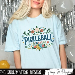 Op de afbeelding: Een lichtblauw t-shirt met een bloemenmotief en de tekst "Pickleball" in blauwe letters.