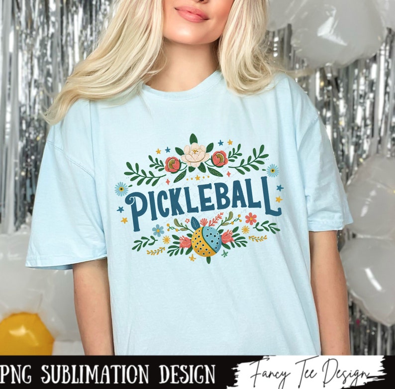 Op de afbeelding: Een lichtblauw t-shirt met een bloemenmotief en de tekst "Pickleball" in blauwe letters.