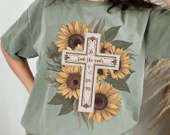 May include: A sublimation design featuring a cross with the text "He took the nails for me" surrounded by sunflowers. The design is on a green t-shirt.