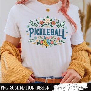 Op de afbeelding: Wit T-shirt met een bloemenmotief en de tekst "Pickleball" in blauwe en gele letters.