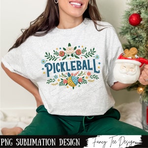 Op de afbeelding: Een wit T-shirt met een bloemenpatroon en de tekst "Pickleball" in blauw. Het ontwerp bevat een pickleball in het midden, omringd door bloemen en bladeren.