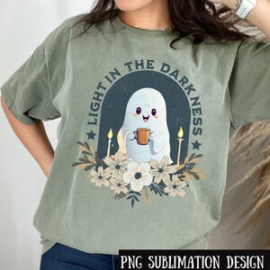 Puede incluir: Una camiseta verde con un fantasma sosteniendo una taza de café y el texto "Light in the Darkness" con detalles florales.