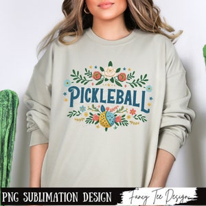 Op de afbeelding: Een lichtgrijs sweatshirt met een bloemenpatroon en de tekst "Pickleball" in blauwe letters. Het ontwerp bevat een pickleball met een bloemenpatroon.
