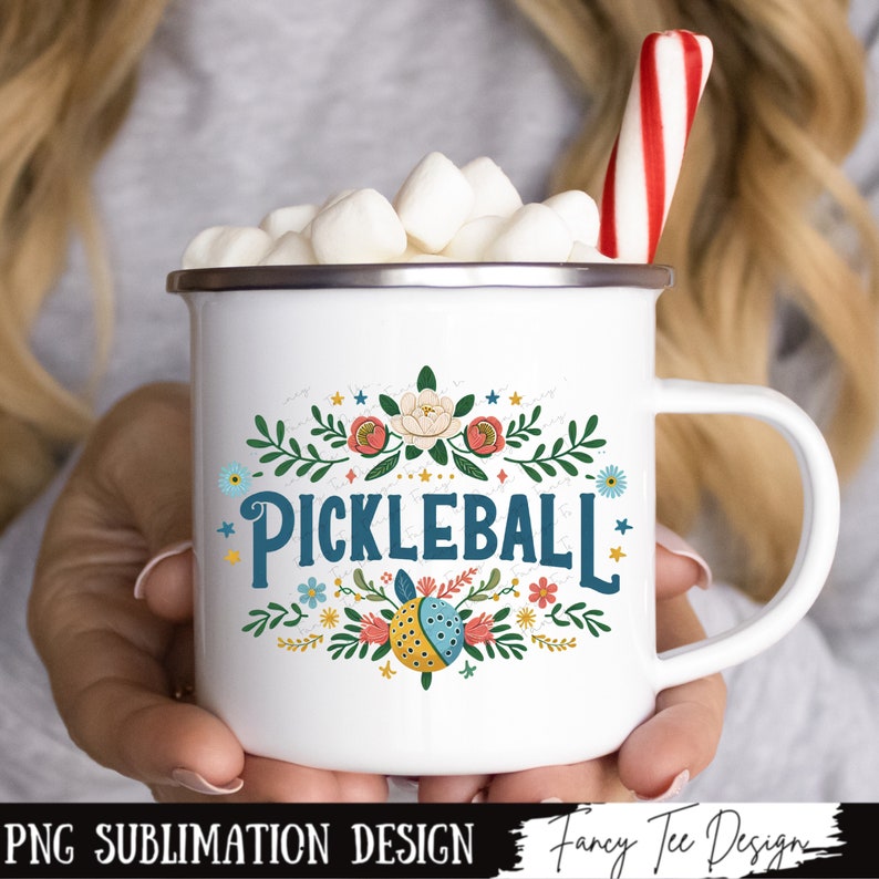 Op de afbeelding: Witte emaille mok met een bloemenmotief en de tekst "Pickleball" in blauw. De mok is gevuld met marshmallows en een rood-witte snoepstok.