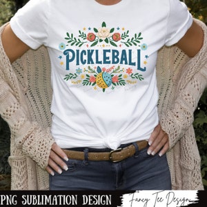 Op de afbeelding: Een wit T-shirt met een bloemenmotief en de tekst "Pickleball" in blauw. Het ontwerp bevat een gele en blauwe pickleball met een lieveheersbeestje erop. De tekst "Family, Friends, Fun" staat in een kleiner lettertype onder de pickleball.