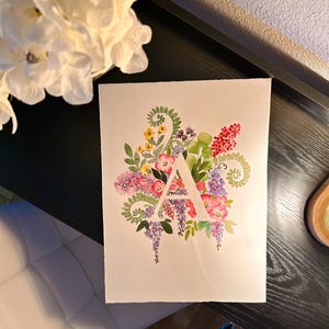 May include: Watercolor painting of the letter A surrounded by a variety of flowers in shades of pink, purple, yellow, and green.