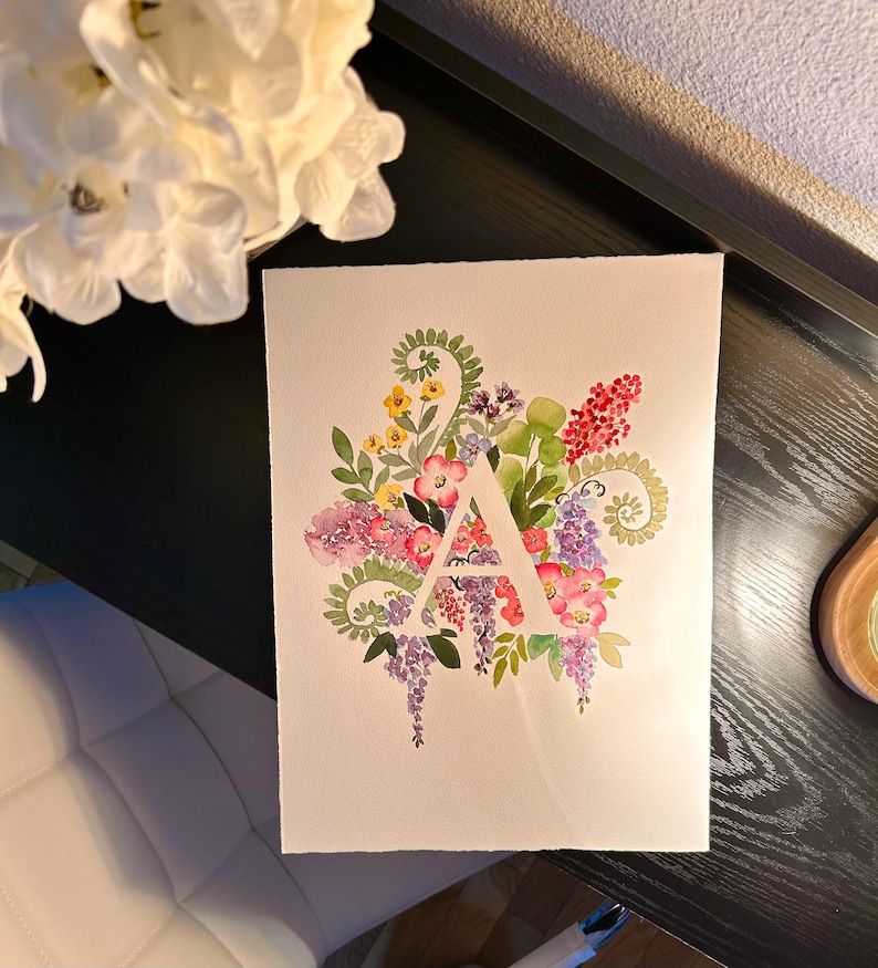 May include: Watercolor painting of the letter A surrounded by a variety of flowers in shades of pink, purple, yellow, and green.