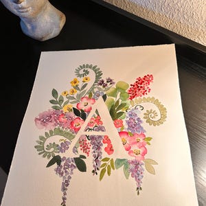 May include: Watercolor painting of the letter A surrounded by a variety of flowers in shades of pink, purple, yellow, and green.