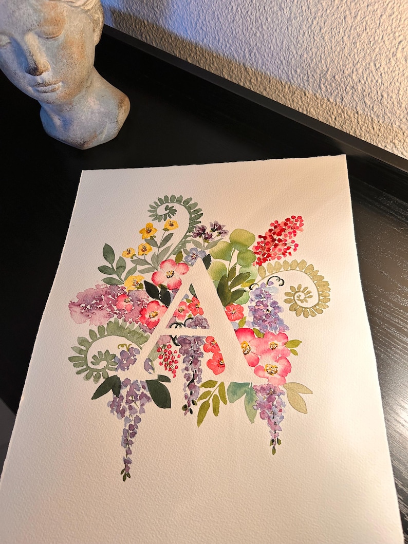 May include: Watercolor painting of the letter A surrounded by a variety of flowers in shades of pink, purple, yellow, and green.