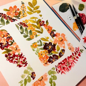 May include: Watercolor floral art on white paper, featuring orange, red, and burgundy flowers with green leaves. A metal watercolor paint set and paintbrushes are visible, suggesting a painting theme.