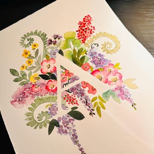 May include: Watercolor painting of the letter A surrounded by a variety of flowers in shades of pink, purple, yellow, and green.