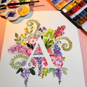 May include: A watercolor painting of the letter A surrounded by a variety of flowers in shades of pink, purple, yellow, and green. The flowers are arranged in a circular pattern around the letter.