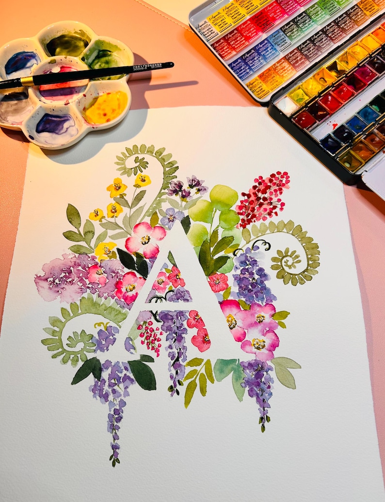 May include: A watercolor painting of the letter A surrounded by a variety of flowers in shades of pink, purple, yellow, and green. The flowers are arranged in a circular pattern around the letter.