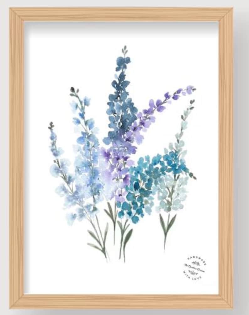 May include: Watercolor painting of blue and purple flowers in a wooden frame. The artwork features a cluster of delicate blooms in shades of blue, teal, and purple against a white background. The frame is a light wood color.