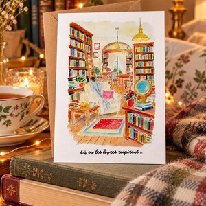 Może przedstawiać: Kartka okolicznościowa z akwarelową ilustracją przytulnej biblioteki z regałami od podłogi do sufitu, wygodnym fotelem, globusem i czerwonym dywanem. Tekst na dole kartki brzmi: "L&agrave; ou les livres respirent..."