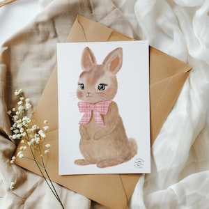 May include: A greeting card with a brown bunny illustration wearing a pink plaid bow tie. The bunny has large eyes and long eyelashes. The card rests on a brown envelope, with a sprig of white flowers in the corner. The overall theme is Easter.
