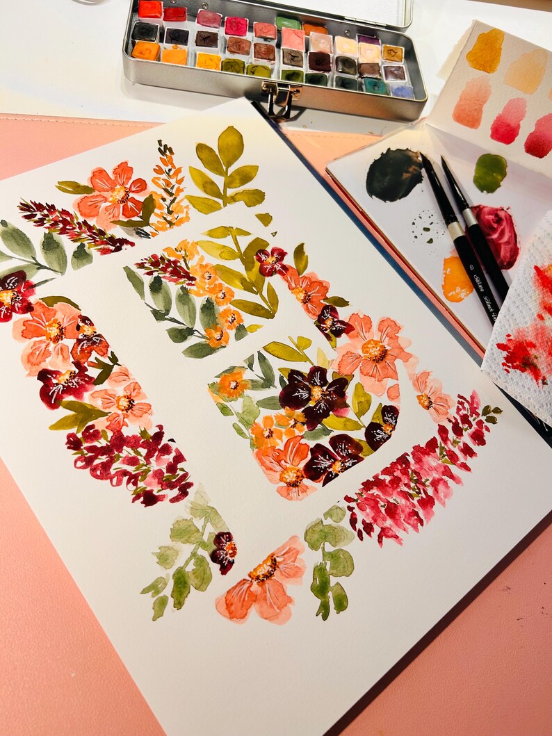 Pu&ograve; includere: Dipinto ad acquerello della lettera "E" decorata con fiori arancioni, rossi e bordeaux e foglie verdi. Una scatola di metallo di acquerelli e una tavolozza con pennelli sono sullo sfondo. L'opera d'arte &egrave; su carta bianca.