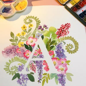 Pode incluir: Pintura em aquarela da letra "A" rodeada por flores coloridas em tons de rosa, roxo, amarelo e verde. Uma paleta de aquarela e um pincel são visíveis no fundo, sugerindo um tema artístico.