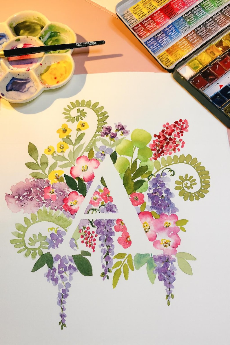 May include: Watercolor painting of the letter "A" surrounded by colorful flowers in shades of pink, purple, yellow, and green. A palette of watercolors and a paintbrush are visible in the background, suggesting an art theme.