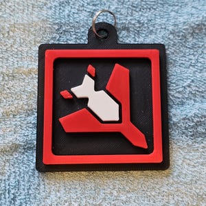 May include: Black, red, and white keychain with a geometric design. The keychain features a black square with a red border and a white and red abstract shape in the center. A silver key ring is attached at the top.