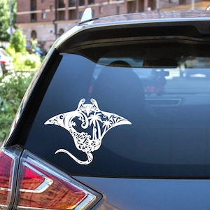 May include: White vinyl decal of a stylised manta ray with palm trees and ocean waves. The decal is on the back window of a silver car.