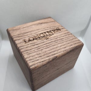 May include: A wooden box with the word "LONGINES" engraved on the top. The box is made of light brown wood with visible grain patterns. The Longines logo is also engraved on the top of the box.