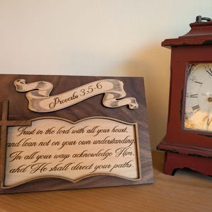 May include: A wooden plaque with the text "Trust in the lord with all your heart" and "Proverbs 3:5-6" is displayed. A cross is attached to the side. A red antique clock is in the background.