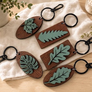 May include: Five keyrings featuring dark brown wooden bases in oval and rectangular shapes. Each keyring is adorned with a raised, light green wooden leaf cutout, including maple, fern, monstera, and oak leaf designs, attached to a black metal ring.