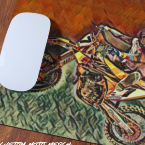 May include: A colorful mousepad featuring a stylized illustration of a motocross rider on a yellow dirt bike. The rider is wearing a blue and orange racing suit and a helmet. The background is a blurred image of a dirt track.